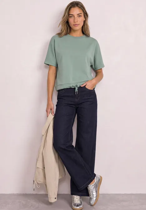 Vrouw in groen T-shirt, donkere wijdvallende jeans en sneakers, met een beige jas in haar hand.