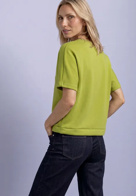 Frau in grüner Kurzarmbluse und dunklen Jeans schaut über ihre Schulter, Hände in den Taschen.