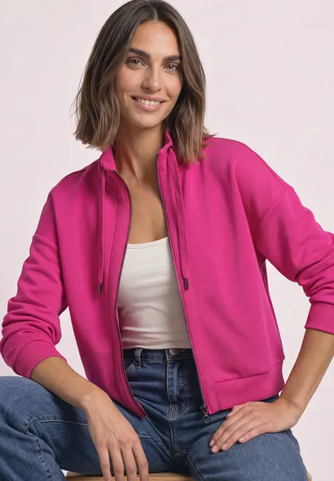 Eine lächelnde Frau in pinkem Kapuzenjacke, weißem Top und Jeans vor hellem Hintergrund.