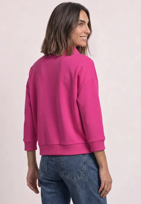 Frau in pinkem Sweatshirt und Jeans, von hinten gezeigt, mit leicht gedrehtem Kopf und Lächeln.