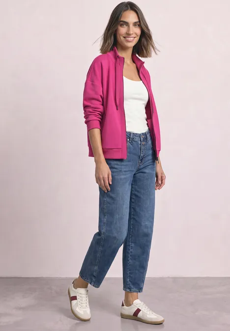 Frau trägt pinke Kapuzenjacke, weißes Shirt, blaue Jeans und weiße Sneaker.