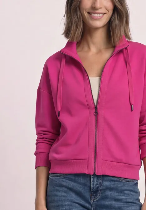 Eine Frau trägt einen pinken, durchgehenden Reißverschluss-Hoodie mit Kapuze und blauen Jeans.