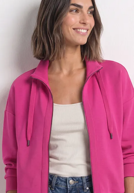 Eine Frau trägt eine pinke Sweatjacke mit Reißverschluss, ein weißes Top und lächelt.