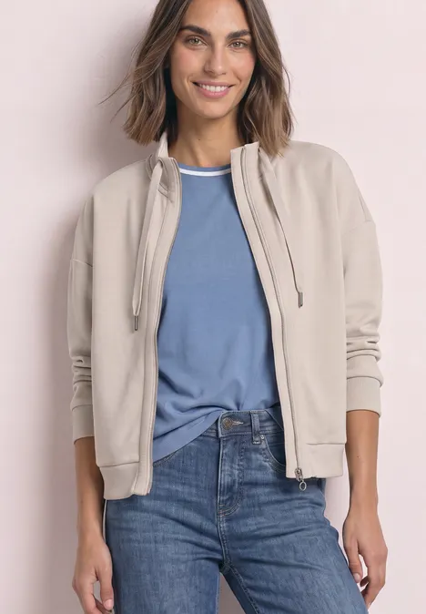 Femme souriante en veste beige, T-shirt bleu et jean, posant devant un fond clair.