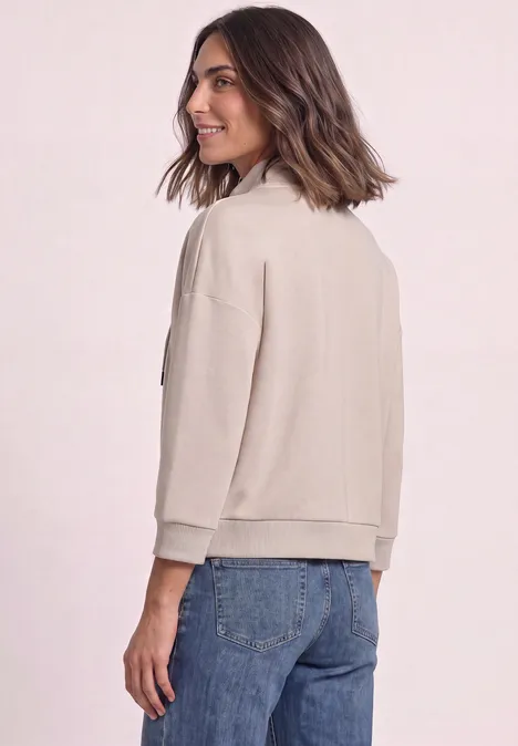 Femme en veste beige et jean, vue de dos en se retournant avec un sourire.