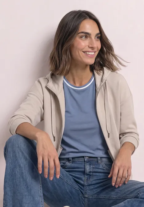 Une femme souriante en jeans, t-shirt bleu et veste à capuche beige, assise devant un fond clair.