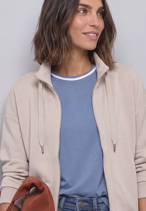 Femme en cardigan beige sur t-shirt bleu, tenant un sac brun, regard de côté.