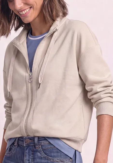 Femme souriante en sweat à capuche beige zippé, t-shirt bleu et jean.
