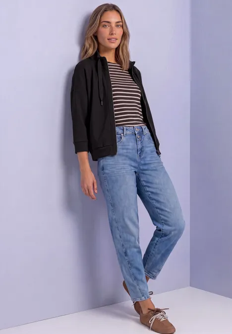 Une femme en veste noire, t-shirt à rayures, jean bleu et mocassins marron devant un mur pastel.