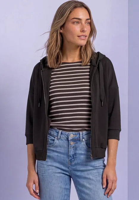 Femme en t-shirt rayé, veste à capuche noire et jean, sur fond clair.