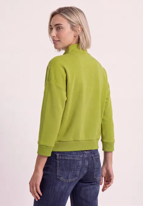 Femme vue de dos portant un pull vert et un jean sur fond clair.