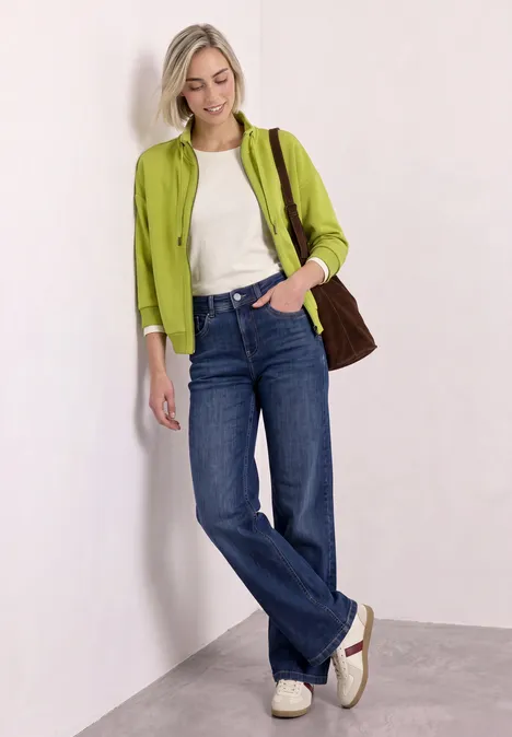 Femme appuyée contre un mur, t-shirt blanc, veste verte, jeans larges, baskets blanches et sac marron.