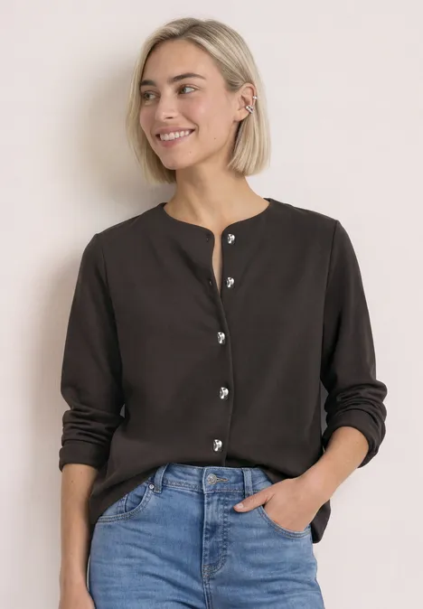 Kobieta z krótkimi blond włosami uśmiecha się, ma brązowy sweter i dżinsy na jasnym tle.