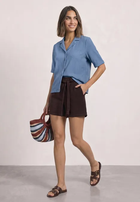 Chemise aux textures variées et à boutons northern blue Frau trägt hellblaues kurzärmliges Hemd, braune Shorts, Sandalen und hält bunte Umhängetasche, lächelnd.