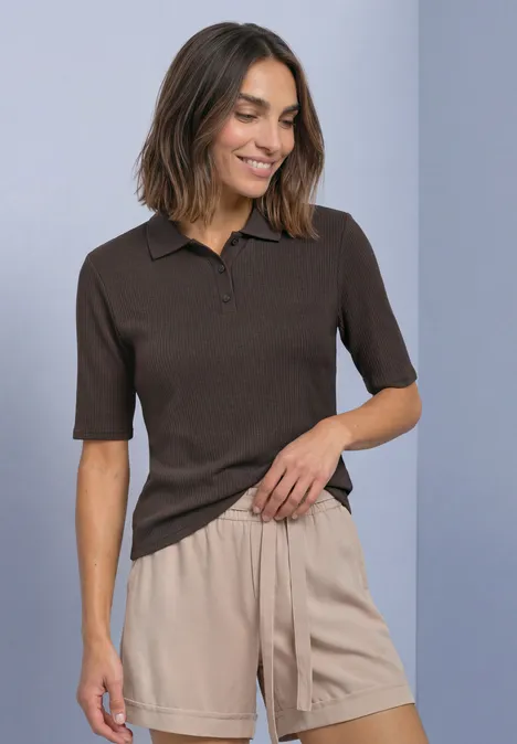 Frau im braunen Kurzarm-Poloshirt und beigefarbenen Shorts, lächelnd, eine Hand in der Tasche.