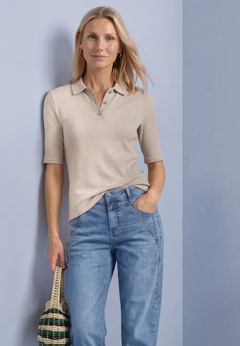 Polo en maille avec motif côtelé clay sand mel. Eine Frau trägt ein beiges Poloshirt und blaue Jeans, hält eine geflochtene Tasche in der Hand.