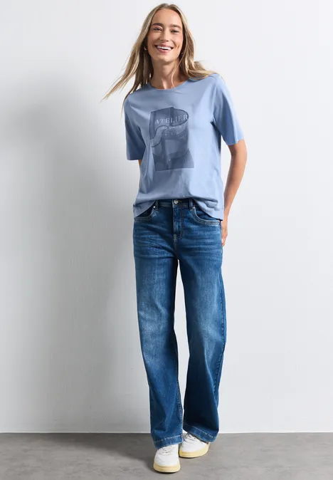T-shirt à col rond avec impression photo northern blue Eine lächelnde Frau in blauem T-Shirt, weiten Jeans und weißen Sneakern vor neutralem Hintergrund.