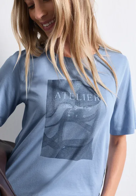 T-shirt à col rond avec impression photo northern blue Eine lächelnde Frau trägt ein hellblaues T-Shirt mit grafischem Print 'Atelier'.