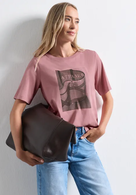 Frau in rosafarbenem T-Shirt mit Print, blauen Jeans und großer brauner Tasche vor weißem Hintergrund.
