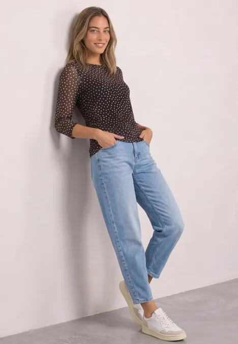 3/4-Arm Shirt aus Mesh mit Print black coffee Frau lehnt an Wand, trägt gepunktetes Top, hellblaue Jeans und weiße Sneakers.