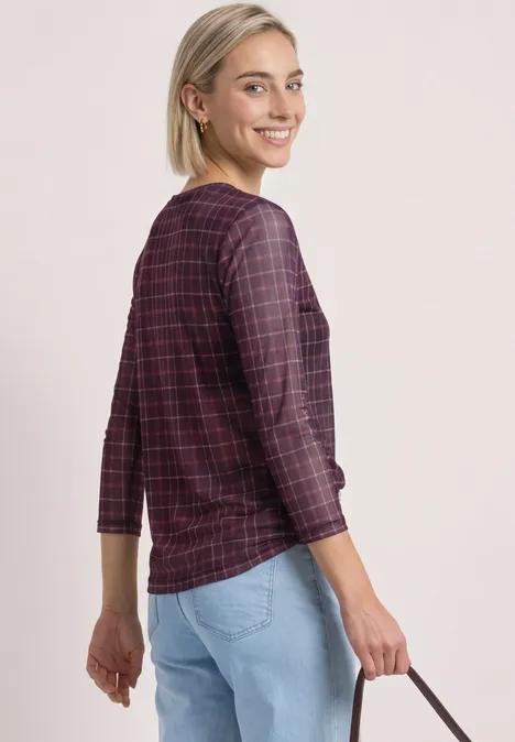 Eine lächelnde Frau in rotem, kariertem Langarmshirt und hellblauen Jeans, die eine kleine Handtasche hält.