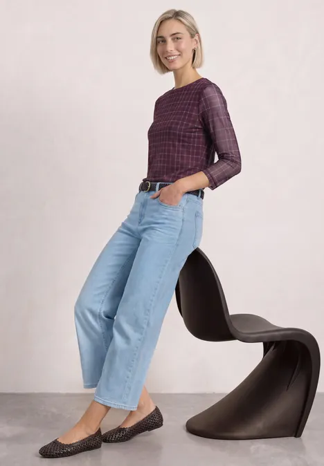 Eine lächelnde Frau in karierter Bluse, lockeren Jeans und schwarzen Ballerinas, lehnt an einem modernen Stuhl.