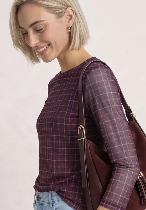 Lächelnde blonde Frau in kariertem violetten Oberteil mit Schultertasche vor hellem Hintergrund.