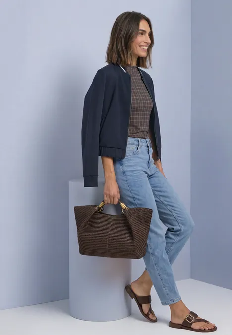 Frau in Jeans, braunem Top und Jacke lehnt lächelnd mit brauner Handtasche vor grauem Hintergrund.