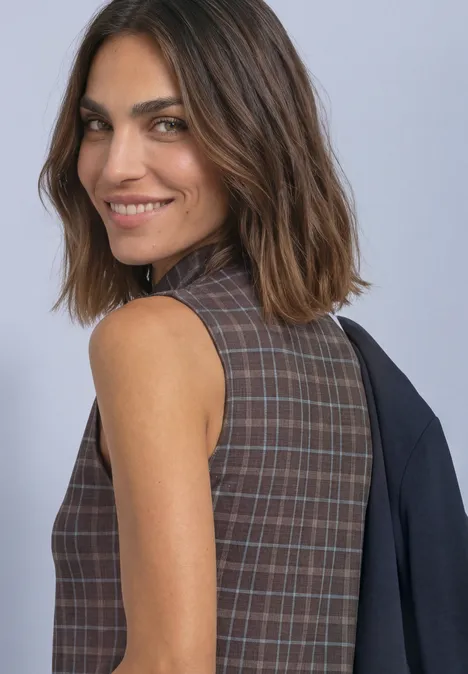 Eine lächelnde Frau in ärmellosem karriertem Kleid legt einen Blazer lässig über die Schulter vor grauem Hintergrund.