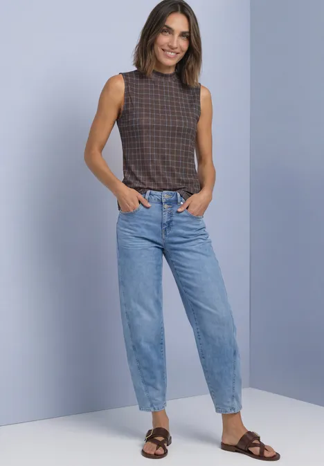Frau steht lächelnd vor hellem Hintergrund, trägt karriertes, ärmelloses Top, lockere Jeans und braune Sandalen.