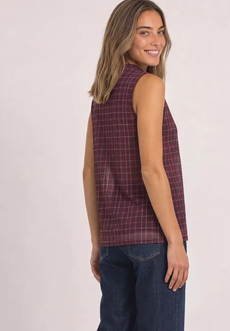 Eine lächelnde Frau in einem ärmellosen karierten Burgunder-Top und blauen Jeans blickt seitlich zur Kamera.