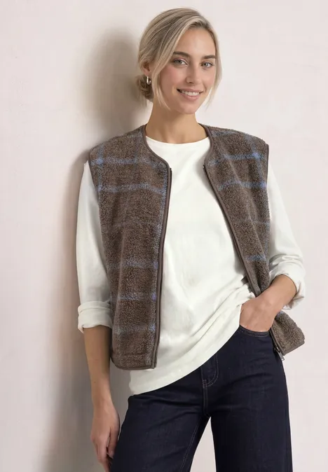 Junge Frau in weißem Langarmshirt, braun-karierter Weste und dunkler Hose, lächelnd vor hellem Hintergrund.