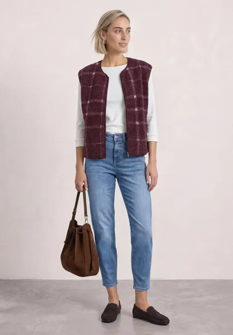 Frau trägt weiße Bluse, burgunderfarben gesteppte Weste, Jeans, schwarze Loafers und braune Tasche.