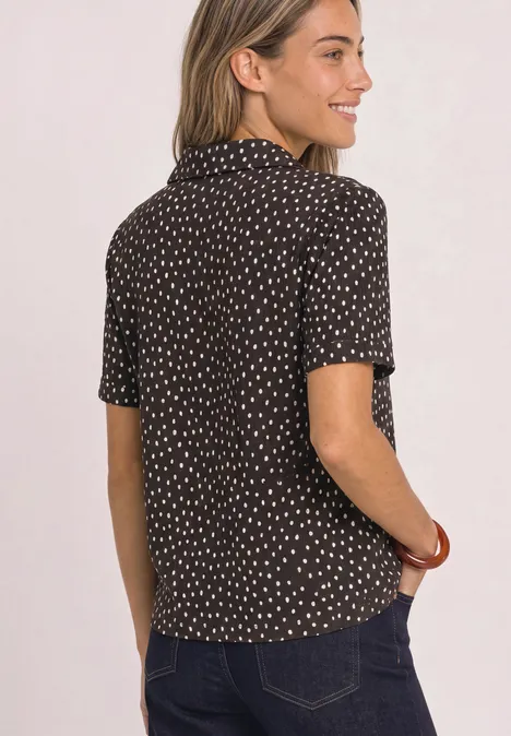 Frau von hinten in schwarzer Bluse mit weißen Punkten und Jeans, die lächelnd zur Seite blickt.