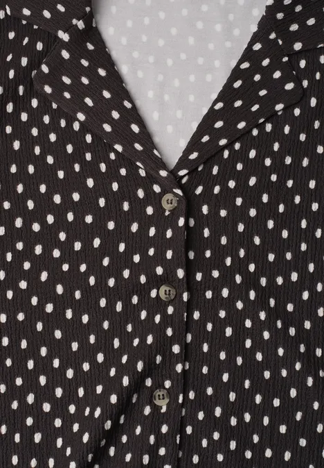 Schwarze Bluse mit weißen Punkten, V-Ausschnitt und drei Knöpfen auf hellem Hintergrund.