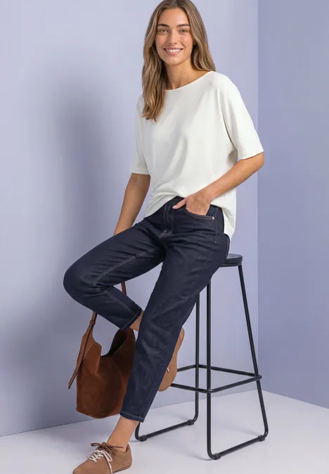 Frau sitzt auf Hocker, trägt weißes T-Shirt, dunkle Jeans und hat eine braune Tasche.