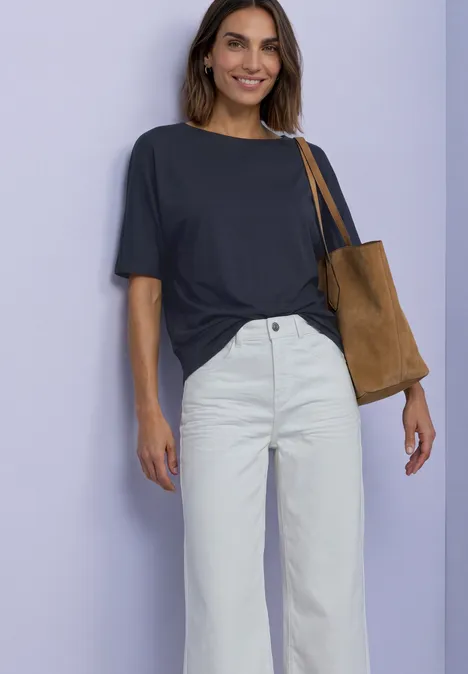 Frau in dunkelblauem T-Shirt, weißer High-Waist-Hose und brauner Schultertasche vor hellem Hintergrund.
