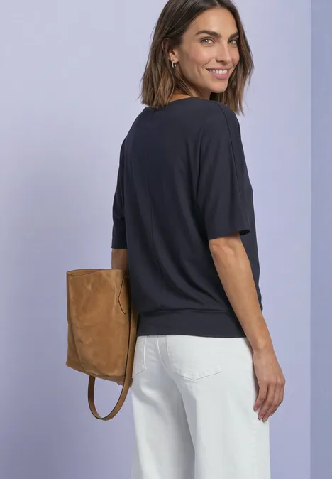 Eine lächelnde Frau in dunkelblauem Shirt und weißen Hosen trägt eine braune Tasche auf dem Rücken.