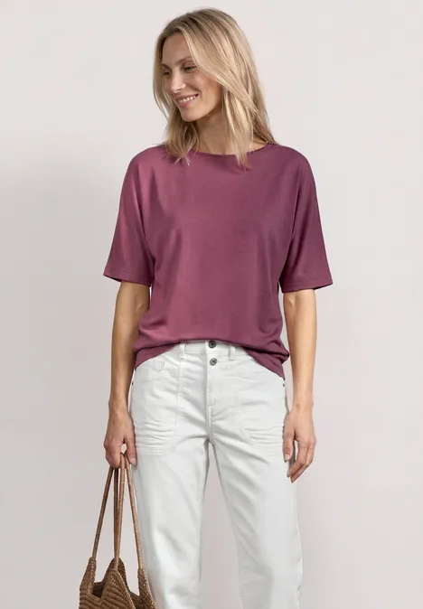 Basic Shirt met boothals en elastische zoom dark clouded blush Een vrouw staat voor een neutrale achtergrond, draagt een paars T-shirt, witte jeans en houdt een bruine tas vast.