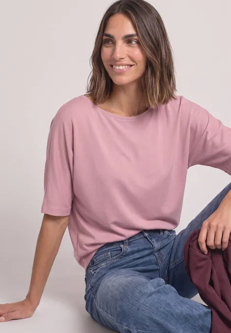Uśmiechnięta kobieta siedzi w różowym t-shircie i dżinsach na neutralnym tle.