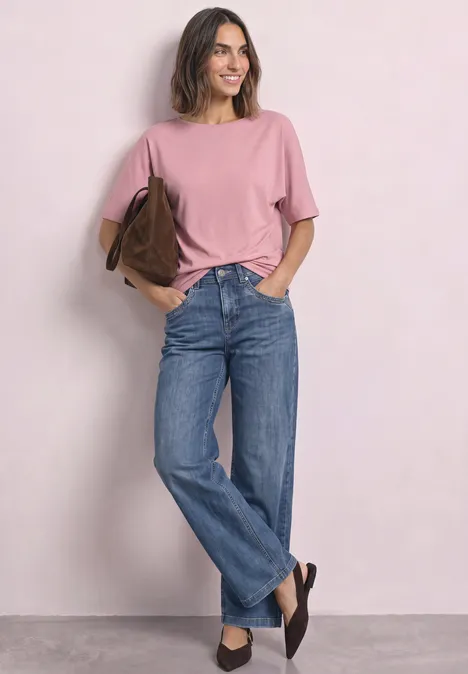 Kobieta w różowym T-shircie, niebieskich jeansach i czarnych butach, uśmiechnięta z brązową torbą na ramieniu.