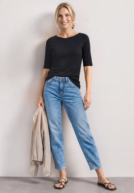 Femme en t-shirt noir, jean taille haute bleu clair, sandales noires, tient une veste beige.
