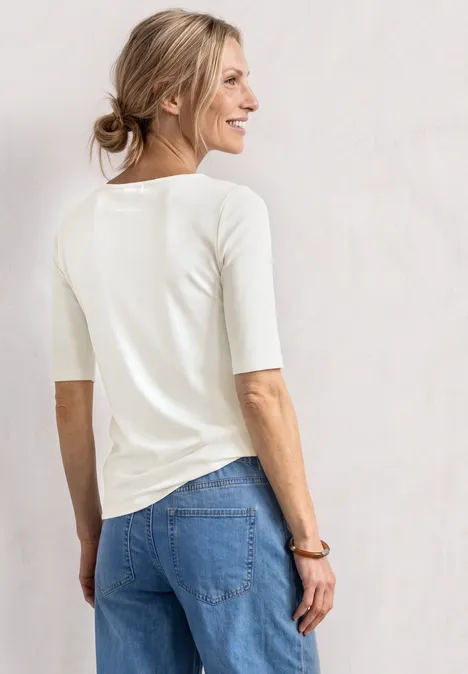 Shirt met golvende halslijn in effen kleur off white Een vrouw staat met haar rug naar de camera, draagt een wit shirt met driekwart mouwen en een blauwe spijkerbroek.