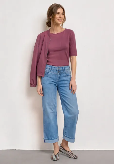 Une femme porte un top et une veste violets sur l'épaule, un jean ample et des slingbacks rayés.