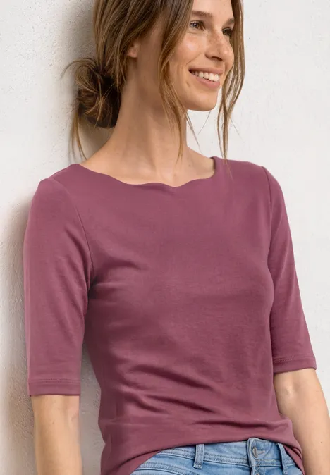 Une femme porte un t-shirt rose à manches mi-longues devant un fond blanc.