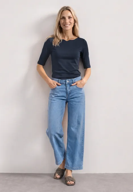 Femme debout devant un mur clair, porte un t-shirt bleu marine manches 3/4, jean large et sandales, mains dans les poches.