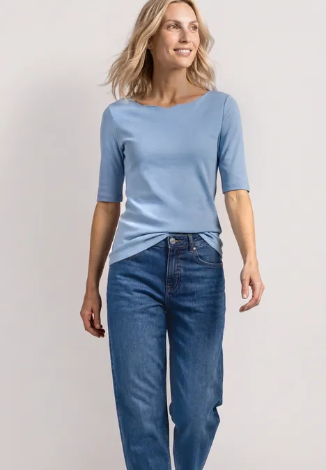 T-shirt à encolure ondulée, couleur unie northern blue Eine Frau steht in einem hellblauen T-Shirt und blauen Jeans vor einem neutralen Hintergrund.