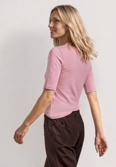 Shirt met golvende halslijn in effen kleur vintage blush Een vrouw van achter gezien draagt een roze shirt met korte mouwen en een donkere broek, lopend opzij.