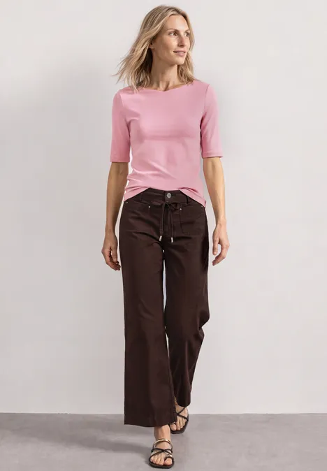 Shirt met golvende halslijn in effen kleur vintage blush Vrouw in roze top en brede bruine broek staat voor een neutrale achtergrond.