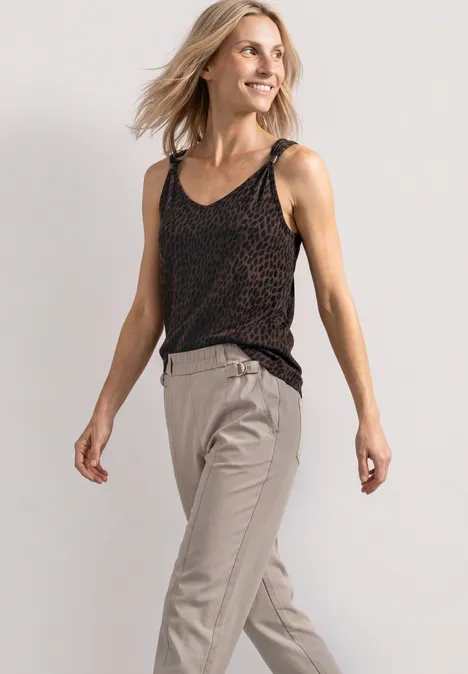 Top met V-hals en decoratief detail molten earth Frau trägt schwarzes gemustertes Top und helle Stoffhose, lächelnd, vor neutralem Hintergrund.